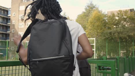 Young-Couple-With-Backpacks-Arriving-At-Artificial-Soccer-Pitch-In-Urban-City-Area-To-Play-Football