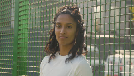 Portrait-Of-Young-Woman-Against-Fence-On-Artificial-Soccer-Pitch-In-Urban-City-Area-