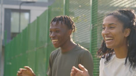 Supporters-Shouting-Encouragement-To-Football-Players-On-Artificial-Soccer-Pitch-In-Urban-City-Area-