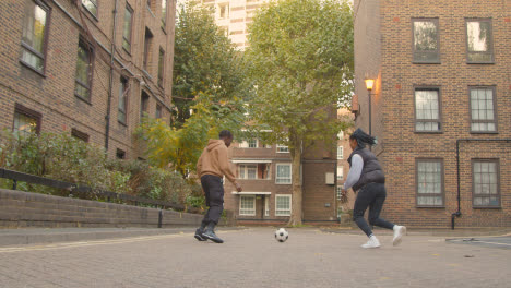 Young-Couple-Kicking-And-Passing-Football-Walking-Along-Street-In-Urban-City-Area-