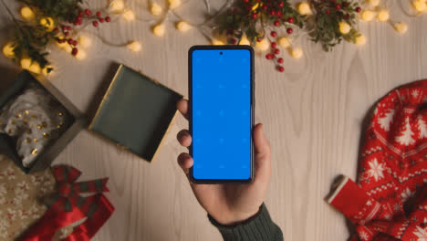 Overhead-Shot-Of-Person-Holding-Blue-Screen-Mobile-Phone-With-Christmas-Decorations-And-Wrapping