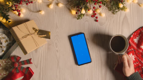 Overhead-Shot-Of-Person-With-Blue-Screen-Mobile-Phone-With-Christmas-Decorations-And-Wrapping