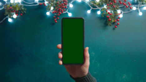 Überkopfaufnahme-Einer-Person,-Die-Ein-Greenscreen-Handy-Mit-Weihnachtsdekorationen-Und-Stechpalmenbeeren-Hält
