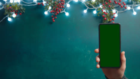 Overhead-Shot-Of-Person-Holding-Green-Screen-Mobile-Phone-With-Christmas-Decorations-And-Holly-Berries-1