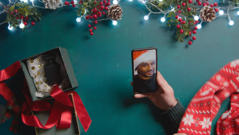 Overhead-Shot-Of-Person-Having-Christmas-Video-Call-With-Friend-On-Mobile-Phone-With-Christmas-Decorations-As-Background