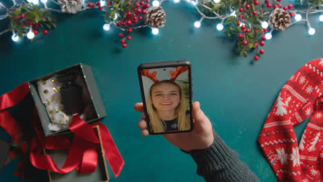 Overhead-Shot-Of-Person-Having-Christmas-Video-Call-With-Friend-On-Mobile-Phone-With-Christmas-Decorations-As-Background-1