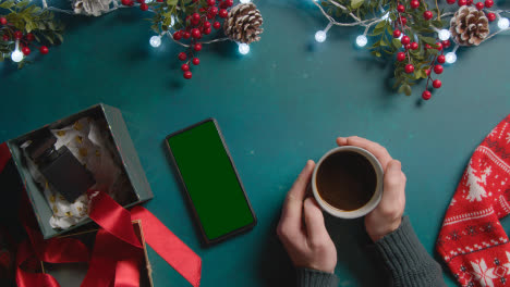 Overhead-Shot-Of-Person-With-Green-Screen-Mobile-Phone-With-Christmas-Decorations-And-Gifts