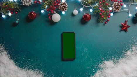 Overhead-Shot-Of-Green-Screen-Mobile-Phone-With-Christmas-Decorations-And-Snow
