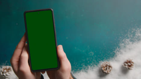 Overhead-Shot-Of-Person-With-Green-Screen-Mobile-Phone-Above-Christmas-Background-1