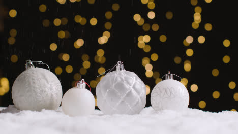 Studio-Christmas-Still-Life-With-Snow-Falling-On-Decorations-With-Lights-On-Background