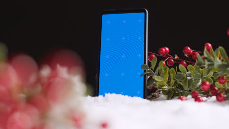 Blue-Screen-Mobile-Phone-On-Christmas-Background-With-Snow-And-Foliage