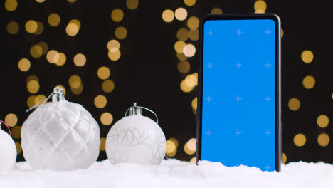 Handy-Mit-Blauem-Bildschirm-Auf-Weihnachtlichem-Hintergrund-Mit-Schnee-Und-Baumschmuck