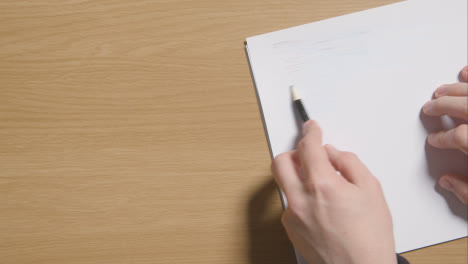 Person-Sketching-With-Pencil-On-Pad-On-Wooden-Table-Background