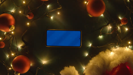 Overhead-Aufnahme-Von-Blue-Screen-Handy-Mit-Weihnachtsschmuck,-Lichtern-Und-Weihnachtsmütze