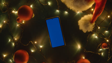 Overhead-Aufnahme-Eines-Sich-Drehenden-Blue-Screen-Handys-Mit-Weihnachtsdekoration,-Lichtern-Und-Weihnachtsmütze
