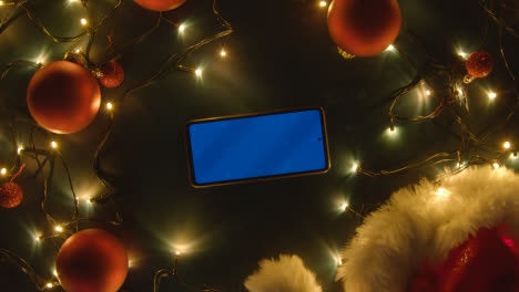 Overhead-Aufnahme-Eines-Rotierenden-Blue-Screen-Handys-Mit-Weihnachtsdekoration,-Lichtern-Und-Weihnachtsmütze-1