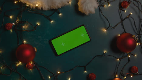 Overhead-Aufnahme-Eines-Rotierenden-Green-Screen-Handys-Mit-Weihnachtsdekoration,-Lichtern-Und-Weihnachtsmütze