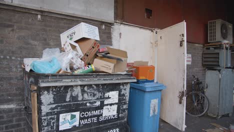 Basura-Desbordante-Y-Contenedores-Con-Ruedas-Delante-De-La-Pared-En-Tower-Hamlets,-Londres,-Gran-Bretaña.