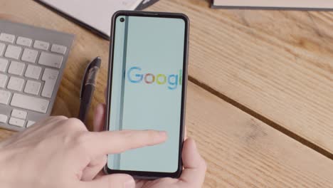 Person-Holding-Mobile-Phone-On-Desk-With-Screen-Showing-Brand-Logos-For-Apple,Google,McDonalds,Microsoft,Tesla,Verizon-And-Visa