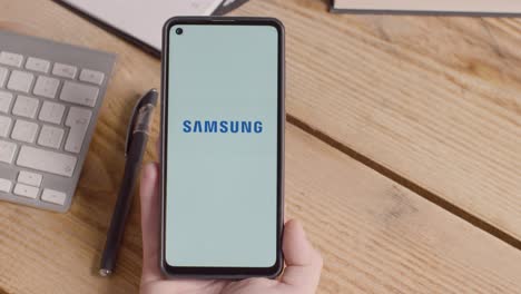 Persona-Que-Tiene-Un-Teléfono-Móvil-En-El-Escritorio-Con-Una-Pantalla-Que-Muestra-Logotipos-De-Marca-Para-Apple-Y-Samsung