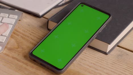 Close-Up-Of-Green-Screen-Mobile-Phone-On-Desk-With-Hand-Typing-On-Computer-Keyboard