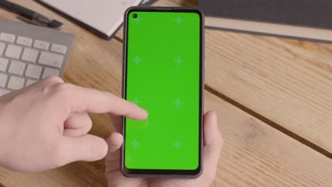 Close-Up-Of-Person-Holding-Green-Screen-Mobile-Phone-On-Desk-With-Hand-Scrolling-Across-Screen