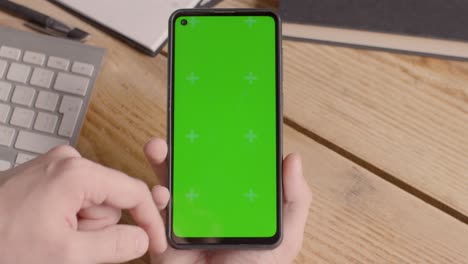 Close-Up-Of-Person-Holding-Green-Screen-Mobile-Phone-On-Desk-With-Hand-Scrolling-Across-Screen-1