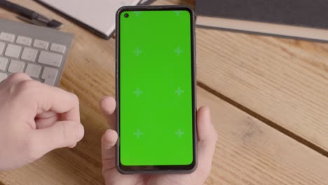 Close-Up-Of-Person-Holding-Green-Screen-Mobile-Phone-On-Desk-With-Hand-Scrolling-Across-Screen-2