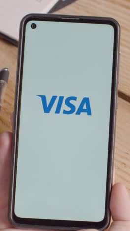 Video-Vertical-De-Una-Persona-Que-Tiene-Un-Teléfono-Móvil-En-El-Escritorio-Con-Una-Pantalla-Que-Muestra-Logotipos-De-Marca-Para-Qualcomm,-Visa,-Mastercard,-American-Express-Y-Paypal