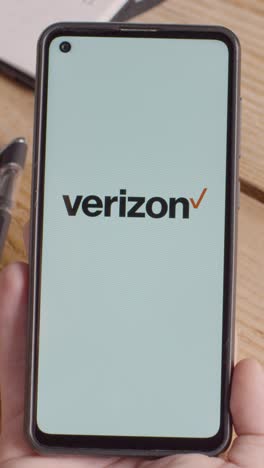 Video-Vertical-De-Una-Persona-Que-Sostiene-Un-Teléfono-Móvil-En-El-Escritorio-Con-Una-Pantalla-Que-Muestra-Los-Logotipos-De-Marca-Para-Tencent,verizon,at