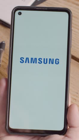 Vertikales-Video-Einer-Hand,-Die-Ein-Mobiltelefon-Auf-Einem-Schreibtisch-Hält,-Mit-Einem-Bildschirm,-Der-Markenlogos-Für-Mastercard,-Samsung,-Tik-Tok,-Alibaba,-Coca-Cola,-Meta-Und-Starbucks-Zeigt