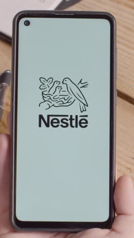 Vertikales-Video-Einer-Person,-Die-Ein-Mobiltelefon-Auf-Einem-Schreibtisch-Hält,-Mit-Einem-Bildschirm,-Auf-Dem-Markenlogos-Für-Mcdonalds,-Nestle,-Coca-Cola,-Starbucks-Und-Pepsico-Zu-Sehen-Sind