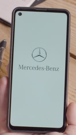 Video-Vertical-De-Una-Persona-Que-Sostiene-Un-Teléfono-Móvil-En-El-Escritorio-Con-Una-Pantalla-Que-Muestra-Logotipos-De-Marca-Para-Tesla,-Mercedes-Benz,-Bmw-Y-Toyota