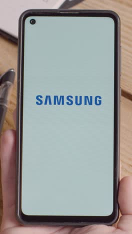 Video-Vertical-De-Una-Persona-Que-Sostiene-Un-Teléfono-Móvil-En-El-Escritorio-Con-Una-Pantalla-Que-Muestra-Logotipos-De-Marca-Para-Apple-Y-Samsung