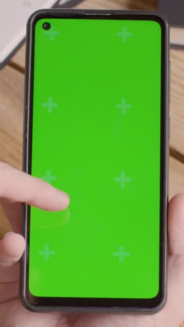 Vertical-Video-Close-Up-Of-Person-Holding-Green-Screen-Mobile-Phone-On-Desk-With-Hand-Scrolling-Across-Screen