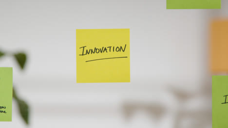 Close-Up-Of-Woman-Putting-Sticky-Note-With-Innovation-Written-On-It-Onto-Transparent-Screen-In-Office