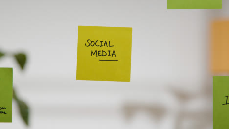 Close-Up-Of-Woman-Putting-Sticky-Note-With-Social-Media-Written-On-It-Onto-Transparent-Screen-In-Office
