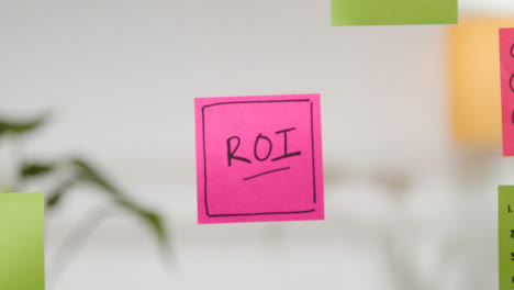 Close-Up-Of-Woman-Putting-Sticky-Note-With-ROI-Written-On-It-Onto-Transparent-Screen-In-Office