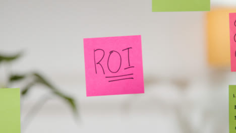 Close-Up-Of-Woman-Writing-ROI-Onto-Sticky-Note-Stuck-To-Transparent-Screen-In-Office