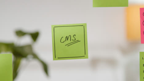 Close-Up-Of-Woman-Putting-Sticky-Note-With-CMS-Written-On-It-Onto-Transparent-Screen-In-Office