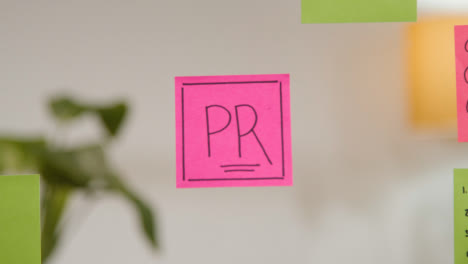 Close-Up-Of-Woman-Writing-PR-Onto-Sticky-Note-Stuck-To-Transparent-Screen-In-Office