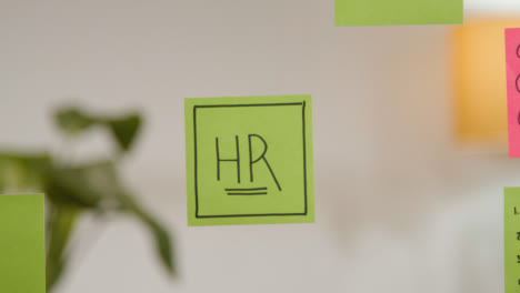 Close-Up-Of-Woman-Putting-Sticky-Note-With-HR-Written-On-It-Onto-Transparent-Screen-In-Office