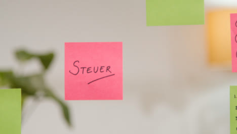 Close-Up-Of-Woman-Putting-Sticky-Note-With-German-Word-Steuer-Or-Tax-Written-On-It-Onto-Transparent-Screen-In-Office-