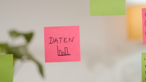 Close-Up-Of-Woman-Putting-Sticky-Note-With-German-Word-Daten-Or-Data-Written-On-It-Onto-Transparent-Screen-In-Office-