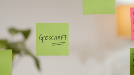 Close-Up-Of-Woman-Putting-Sticky-Note-With-German-Word-Geschaft-Or-Shop-Written-On-It-Onto-Transparent-Screen-In-Office-