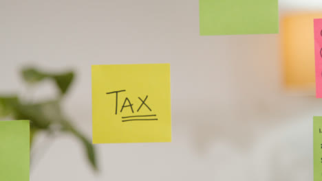 Close-Up-Of-Woman-Putting-Sticky-Note-With-Tax-Written-On-It-Onto-Transparent-Screen-In-Office-