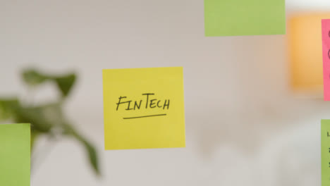 Close-Up-Of-Woman-Putting-Sticky-Note-With-Fintech-Written-On-It-Onto-Transparent-Screen-In-Office-
