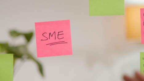 Close-Up-Of-Woman-Putting-Sticky-Note-With-SME-Written-On-It-Onto-Transparent-Screen-In-Office-