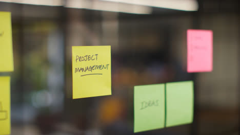 Close-Up-Of-Woman-Putting-Sticky-Note-With-Project-Management-Written-On-It-Onto-Transparent-Screen-In-Office-