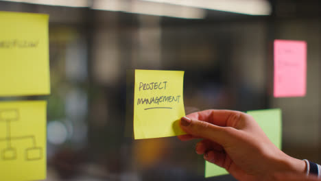 Close-Up-Of-Woman-Putting-Sticky-Note-With-Project-Management-Written-On-It-Onto-Transparent-Screen-In-Office-1
